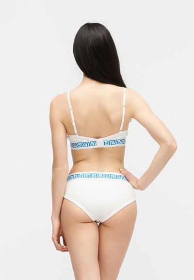 Γυναίκα με μακριά σκούρα μαλλιά φοράει σετ λευκό σουτιέν και εσώρουχο με μπλε μοτίβο ελληνικό κλειδί, στέκεται με το ένα χέρι στη μέση, με την πλάτη προς τα εμπρός.