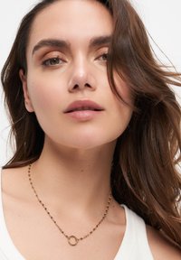 Goldene Kettenhalskette mit rundem Anhänger, bestehend aus verknüpften Segmenten und einer polierten Oberfläche; getragen mit einem schlichten weißen Oberteil.