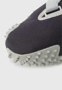 Sportschuh mit einer strukturierten dunkelgrauen Oberseite, grauen Gummispikes auf der Sohle für besseren Halt und einem strukturierten Riemendetail.