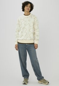 Jeune personne debout, portant un pull crème orné de grandes fleurs marron en contour, un jean bleu et des baskets beiges sur un fond clair uni.
