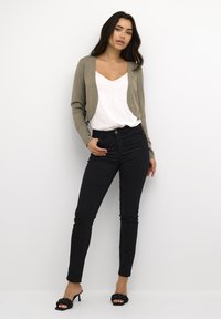 Hellbraunes, kurz geschnittenes Strickjacke über einem weißen V-Ausschnitt-Top, kombiniert mit schwarzen Skinny-Jeans und schwarzen geflochtenen Absatzsandalen. Glatte Texturen, lässige Passform.