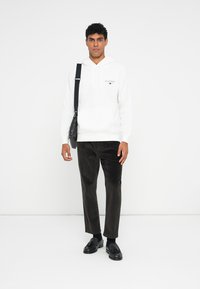 Manta de algodão branca com bolso frontal, apresentando um pequeno logótipo. Combinada com calças de veludo cotelê escuras e sapatos de pele pretos.