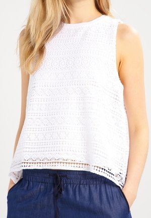 Mujer con top blanco sin mangas con patrón de encaje combinado con pantalones azul marino con cordón, manos casualmente en los bolsillos.