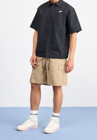 Person som bär en svart kortärmad skjorta med knäppning, beige cargoshorts, vita hög-top sneakers och vita strumpor med svarta ränder, stående på en blå golv.
