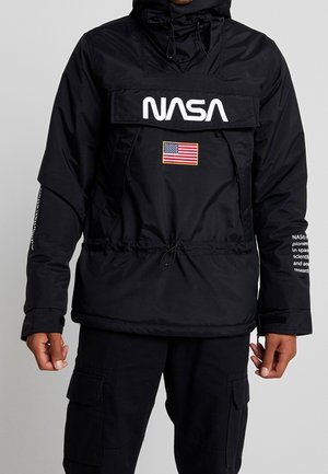 Person iført en sort NASA-jakke med en frontklap, et amerikansk flagpatch under logoet og hvid tekst på højre ærme.