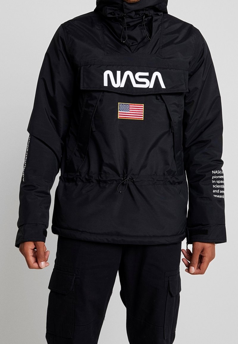 Personne portant une veste noire NASA avec un rabat avant, un écusson du drapeau américain sous le logo, et un texte blanc sur la manche droite.