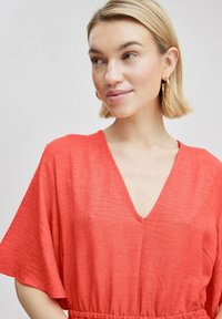 Korallefarbener V-Ausschnitt-Jumpsuit mit kurzen, weiten Ärmeln, gerafftem Bund und einem strukturierten Stoff. Goldene Creolen ergänzen das Outfit.