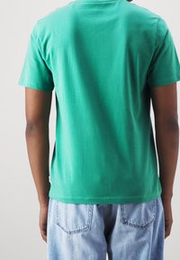 Camiseta de algodón verde con cuello redondo, mangas cortas y dobladillo recto. Combinada con jeans de mezclilla azul claro, mostrando un ajuste casual.