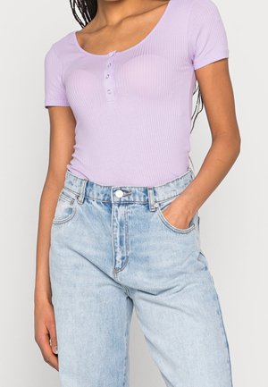 Vrouw draagt een geribd lila topje met korte mouwen en knopen, gecombineerd met een lichtblauwe denim jeans met hoge taille, één hand in haar zak, tegen een effen achtergrond.