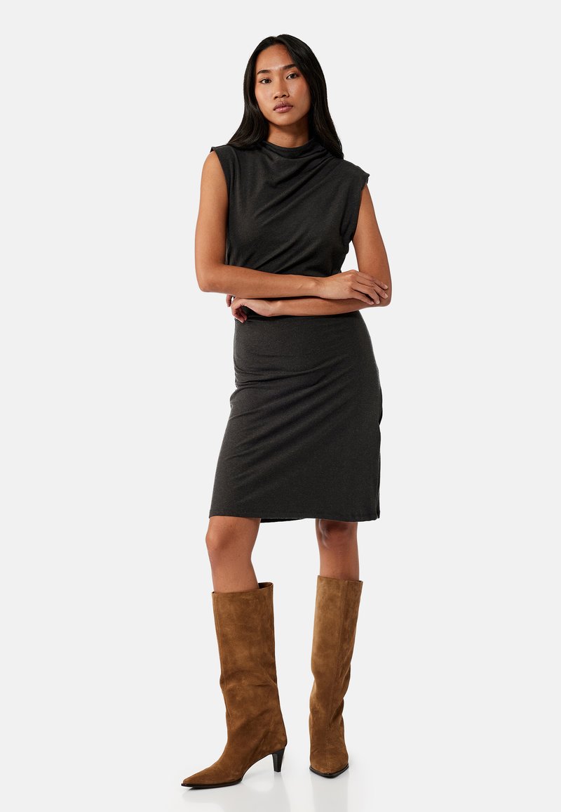 Mujer con vestido negro sin mangas hasta la rodilla y botas marrones de ante hasta la rodilla, de pie con los brazos cruzados y expresión neutral.