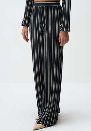 Vrouw draagt een zwarte broek met hoge taille met verticale groene, roze en witte strepen, gecombineerd met beige puntige schoenen.