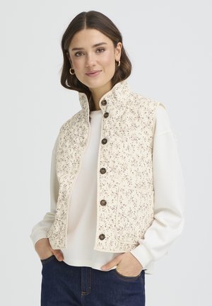 Ženska z rjavimi lasmi nosi kremasto prešito telovnik s cvetličnim vzorcem in velikimi gumbi čez belo majico z dolgimi rokavi ter temno modre kavbojke, z rokami v žepih.