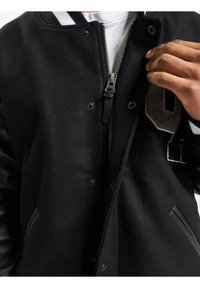 Veste varsity noire avec un corps en laine, des manches en cuir et un zip sur le devant. Équipée de boutons-pression et d'un numéro brodé sur la poitrine.