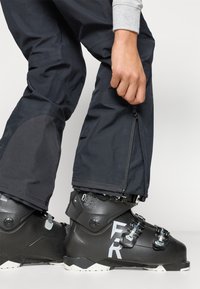 Botas de esqui pretas com fechos de metal e uma superfície texturizada; combinadas com calças escuras que apresentam uma abertura com zíper no tornozelo.