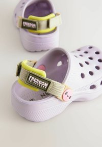 Zoccoli viola con cinturino regolabile giallo e beige. Presentano un'etichetta "LIBERTÀ DI ESPLORARE" e un pulsante sorridente rosa. Perforati per la traspirabilità.
