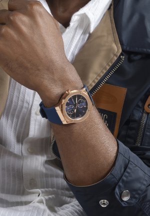 Man wearing a rose gold and blue wristwatch with two dials, dressed in a white shirt and a blue and beige jacket.