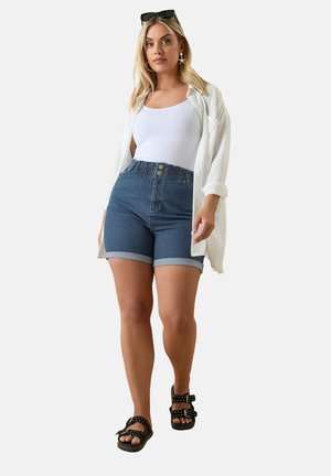 Shorts de denim con dobladillos enrollados y una cintura fruncida, combinados con una camiseta sin mangas blanca y una camisa ligera blanca, llevados con sandalias negras.