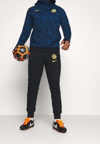 Tenue de sport Nike comprenant un sweat à capuche bleu à motif, un pantalon de survêtement noir avec logo, et des chaussures de football noires avec des accents orange, tenant un ballon.