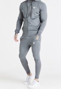 Survêtement de sport gris en tissu lisse, comprenant une veste zippée avec un logo sur la poitrine et un pantalon ajusté avec des poches latérales.