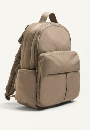 Beige nylon rygsæk med en rund form, polstret design, frontlomme med lynlås og justerbare stropper. Blød tekstur og forstærkede sømme er synlige.