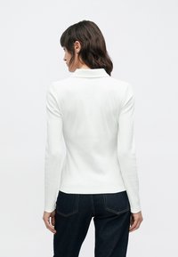 Biela polokošeľa s dlhým rukávom so žíhanou textúrou a klasickým golierom. Nosí sa s tmavomodrými džínami, ktoré majú viditeľné švy.