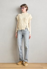 Top a maniche corte di colore crema lavorato a maglia con design a diamante e lacci laterali, abbinato a jeans dritti azzurri e scarpe con piattaforma color tan.