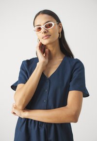 Navyblaues Leinenkleid mit kurzen Puffärmeln und V-Ausschnitt, ausgestattet mit einer durchgehenden Knopfleiste und einer taillierten Taille. Weiße Sonnenbrille mit braunen Gläsern.