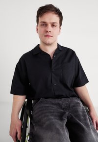 Kortærmet sort skjorte med knapper og et brystlomme, parret med grå denim jeans. Individ i en kørestol.