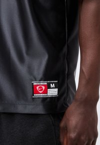 Κοντινό πλάνο ενός σκουρόχρωμου μπράτσου δίπλα στο κάτω μέρος μιας μαύρης φανέλας ποδοσφαίρου Nike με κόκκινη και λευκή ετικέτα μεγέθους M.