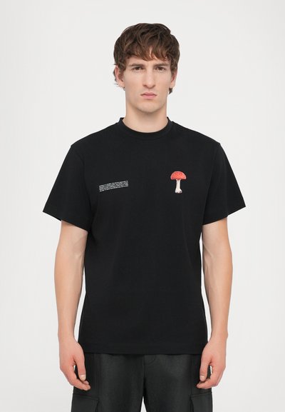 T-shirt en coton noir avec un petit graphique de champignon rouge et blanc sur la poitrine et un petit texte sur le côté gauche. Design à col rond.
