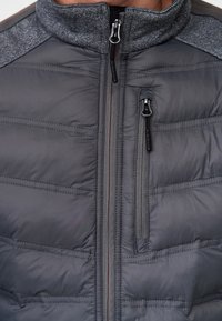 Nahaufnahme einer grauen, gesteppte Jacke mit hohem Kragen, zentralem Reißverschluss und einer senkrechten, mit Reißverschluss verschließbaren Brusttasche auf der rechten Seite.