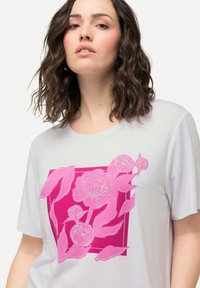 Camiseta de manga corta blanca con un gran gráfico floral en tonos rosas y un detalle intrincado, sobre un fondo cuadrado rosa.