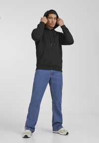 Black hoodie with a front pocket and adjustable drawstrings, paired with blue denim jeans and beige athletic shoes on a neutral background.