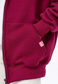 Bordová mikina na zip s klokaní kapsou, žebrovanými manžetami a malou růžovou visačkou na kapse. Měkká, strukturovaná bavlněná směs.