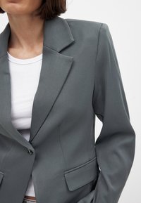Grå blazer med klapper, enkelt knaplukning, to forlommer og glat tekstur, båret over en hvid ribbet tanktop.