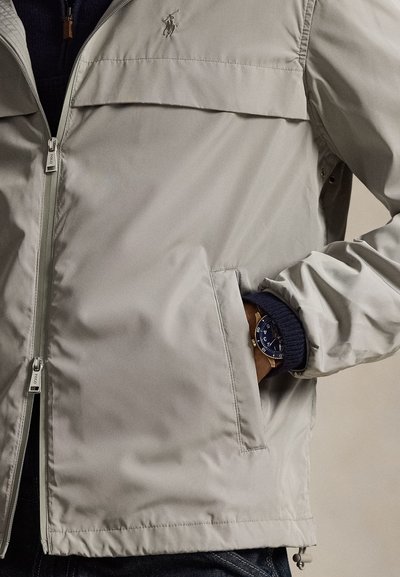 Ελαφρύ γκρι μπουφάν με φερμουάρ, πλαϊνή τσέπη και έμβλημα. Σημαντικά χαρακτηριστικά είναι η ριμπ πλεκτή μανσέτα σε ναυτικό μπλε και ένα ρολόι χειρός.