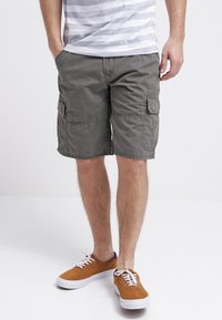 Shorts cargo color khaki con molte tasche laterali, materiale in cotone, vestibilità rilassata e orlo sopra il ginocchio, abbinati a sneakers in suede marrone.