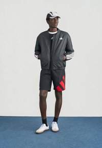 Zwarte bomberjack met witte strepen, zwarte sportshorts met rode accenten, witte sneakers en een grijze pet. Eenvoudig, sportief ontwerp.