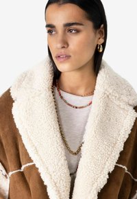 Manteau en daim marron avec un col blanc et duveteux, porté sur une chemise gris clair. Les bijoux en couches notables comprennent des perles colorées et une chaîne en or.