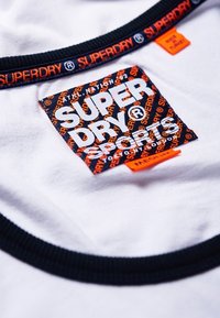 Camiseta blanca con cuello redondo en azul marino. Presenta una etiqueta de marca colorida con "SUPERDRY" en letras grandes y una etiqueta de acento naranja en el lateral.