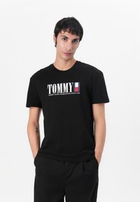 Sort T-skjorte med ordet "TOMMY" i fete hvite bokstaver, med en logoakcent. Korte ermer og rund hals. Laget av bomull.