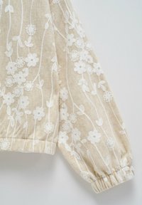 Tissu beige avec broderie florale blanche, manches longues bouffantes avec poignet élastique.