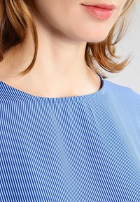 Žena se světlou pletí a světle hnědými vlasy, oblečená v modro-bíle pruhované košili, ukazuje krk a dolní část obličeje.