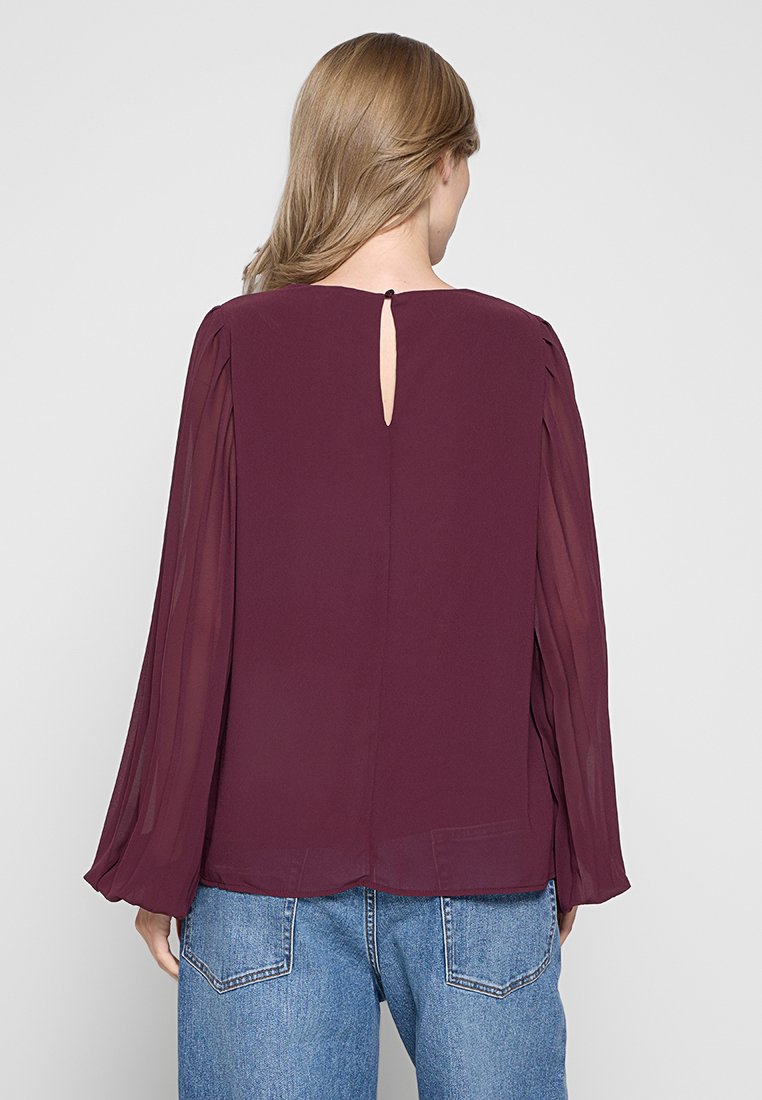 Burgunderbluse mit transparenten langen Ärmeln, rückseitigem Schlitzdetail und lockerer Passform, kombiniert mit hoch taillierten blauen Jeans.