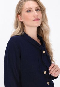 Navy cardigan made of soft fabric featuring a shawl collar, gold buttons, and a tie detail. Smooth texture with a relaxed fit.