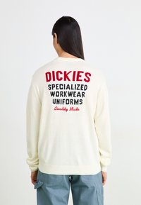 Pull en tricot crème avec du texte rouge et noir dans le dos : "UNIFORMES DE TRAVAIL SPÉCIALISÉS DICKIES, qualité supérieure." Poignets côtelés.
