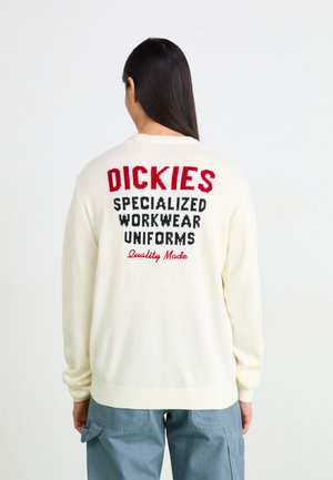 Kremasti pleteni džemper s crvenim i crnim tekstom na leđima: "DICKIES SPECIALIZED WORKWEAR UNIFORMS Quality Made." Ribljenkasti manžeti.