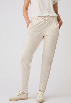 Vrouw draagt beige joggingbroek met koord en zakken en witte sneakers, staand tegen een effen achtergrond.