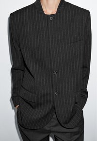 Osoba v tmavém pruhovaném saku s vysokým límcem, se třemi zapnutými knoflíky, s rukama v kapsách, na jednoduchém světlém pozadí.