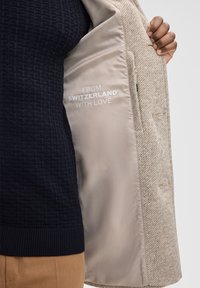 Beige textuurjas met een lichte satijnen voering, met "FROM SWITZERLAND WITH LOVE" in het wit gedrukt. Ribbelige marineblauwe trui eronder.